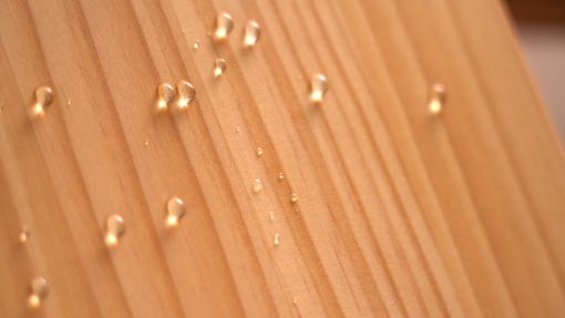 塗裝有撥水劑的木板以傾斜角度立起，數顆水珠從上而下沿木板滑落無滲入情形