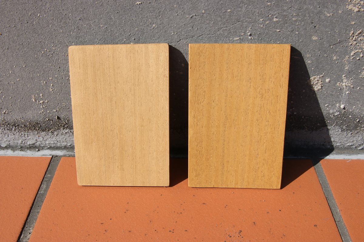 兩片顏色相近的木板倚靠水泥牆邊立起，放置在戶外的陽光下。左方木板較右方木板色彩淺
